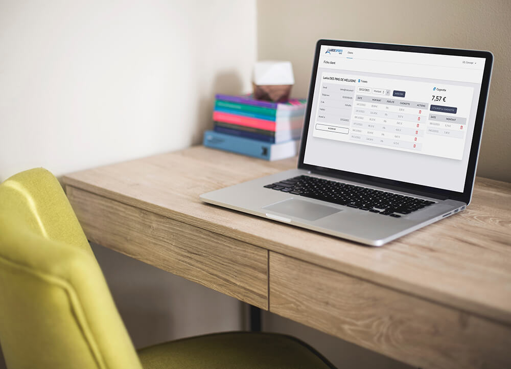 On voit un bureau avec un MacBook gris posé dessus. L'écran affiche un application réalisée par J2L Concept. 