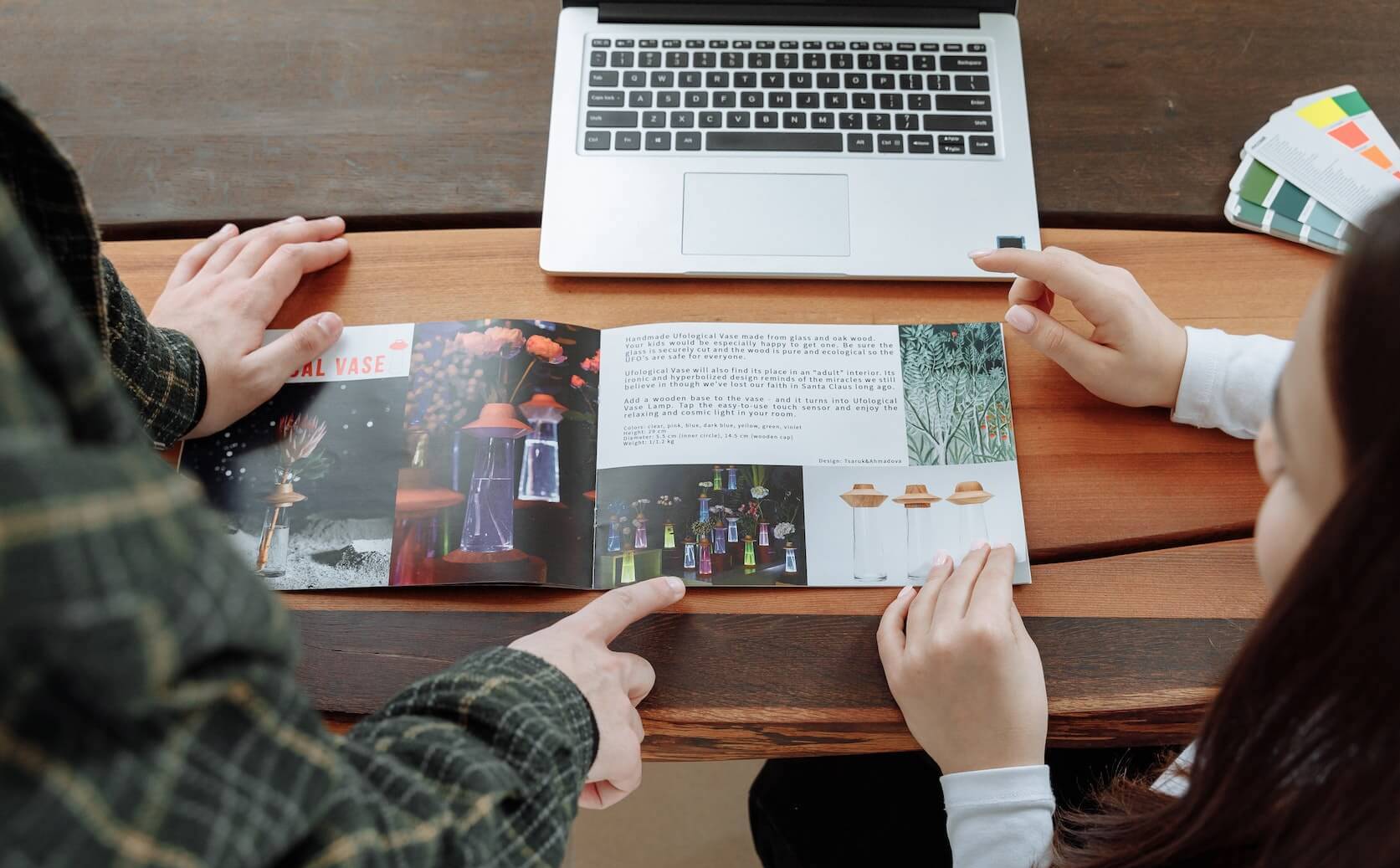 On voit deux personnes penchées sur un bureau. Il y a un prospectus et un MacBook posé sur ce bureau. 